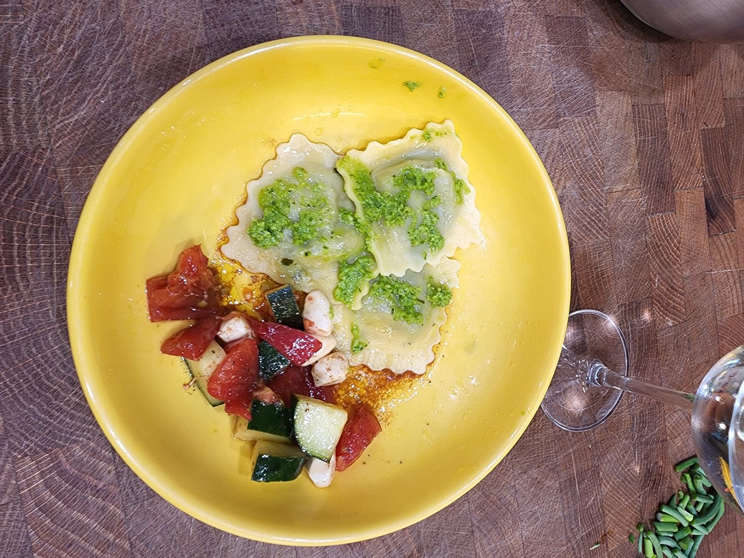 Plate of homemade ravioli topped with green pesto, served alongside a fresh salad of diced tomatoes, zucchini, and mozzarella, with a glass of water on a wooden table.