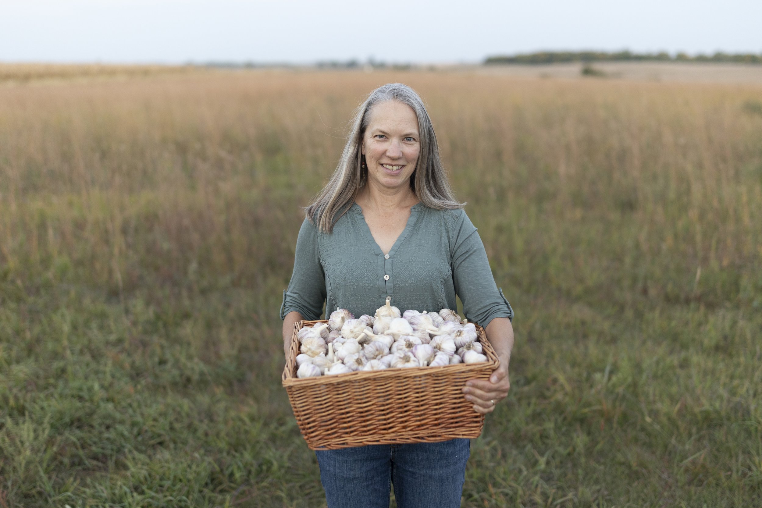 Woman holding a large basket of freshly harvested garlic, standing in a grassy field with a scenic background. The image showcases the beauty of agricultural farming and the joy of harvesting organic produce.
