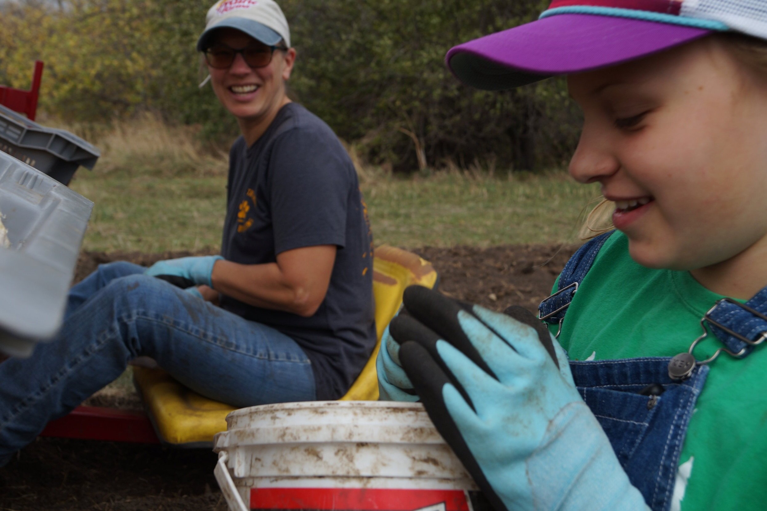 A woman and a girl are enjoying a gardening activity outdoors. The woman, wearing glasses and a cap, smiles while sitting on a yellow seat. The girl, dressed in a green shirt and denim overalls, is focused on her task, wearing gloves and holding a small object. A dirty bucket is visible nearby, indicating they are engaged in planting or harvesting.