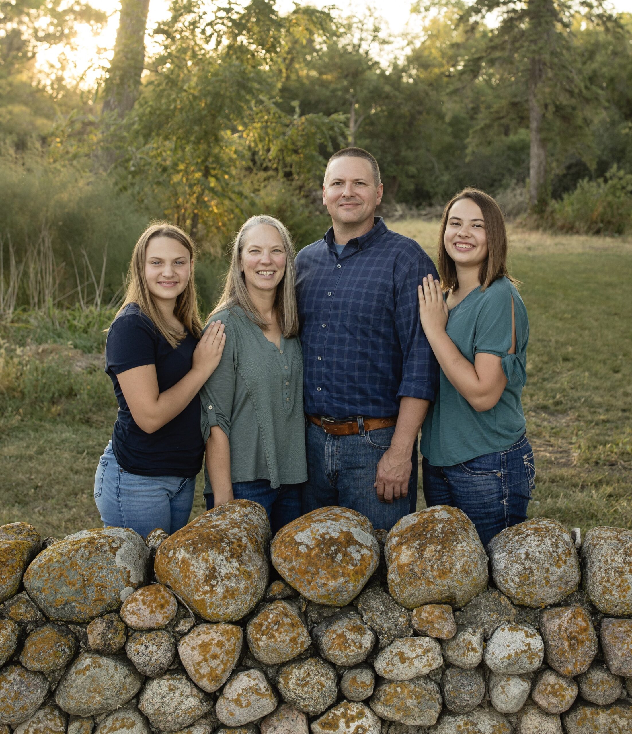 Family portrait featuring four members standing together by a stone wall in a natural outdoor setting, with trees and soft sunlight in the background. The group consists of two teenage daughters, a mother, and a father, all smiling and dressed casually.